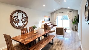 Un comedor con una mesa y bancos de madera, un espejo grande y vistas a una sala de estar con un sofá de cuero y una planta.