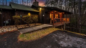 A rustic cabin with a porch and a stone chimney, surrounded by trees.