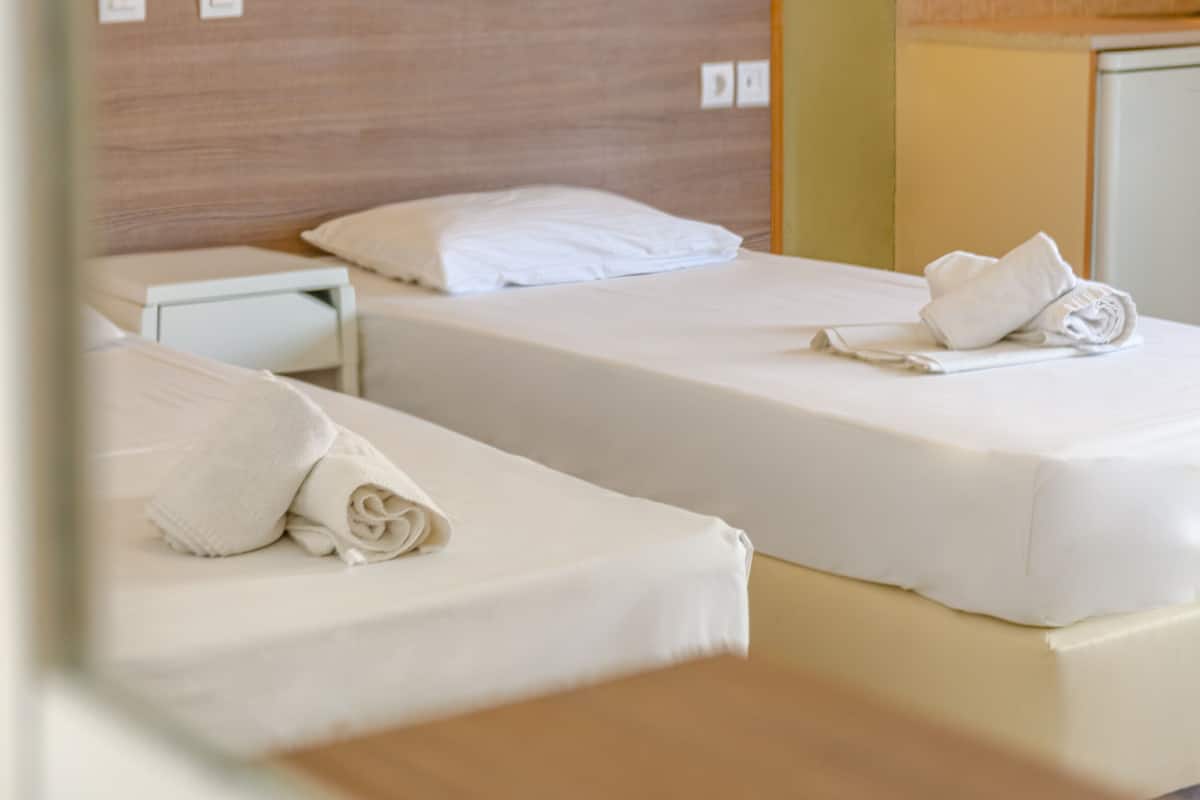 A hotel room with two single beds, each with a white mattress and pillow, and a neatly folded towel on the bed.