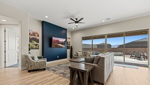 A spacious living room with a large window, a ceiling fan, and a flat-screen TV mounted on the wall.
