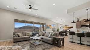 A spacious living room with a large sofa set, a coffee table, and a dining area with a kitchen in the background.