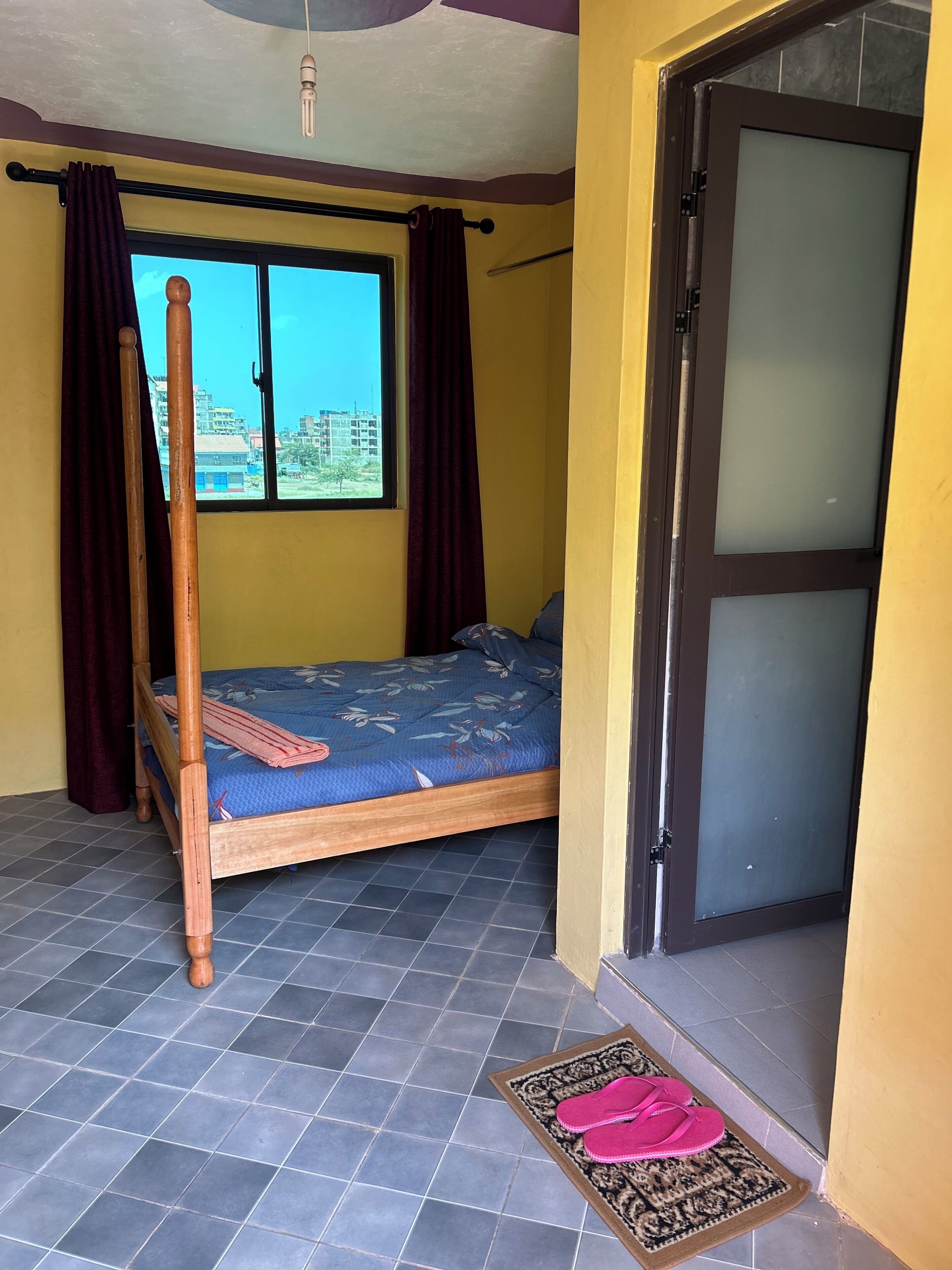 A single bed with a blue mattress and wooden frame, a window with curtains, a door with a frosted glass panel, and a tiled floor.