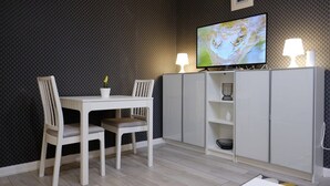A dining area with a white table and chairs, a flat-screen TV mounted above, and a cabinet with shelves below. - Joy City Stay Versului 8 (Timișoara)