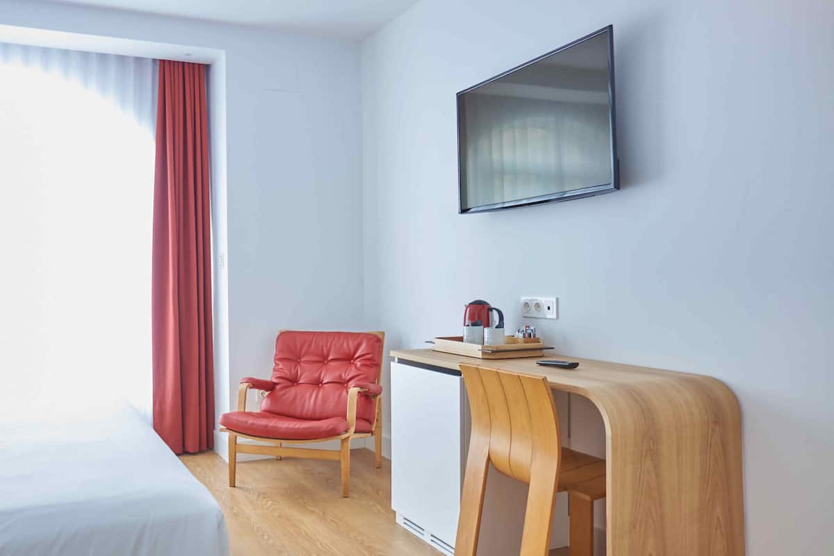 A hotel room with a bed, a desk with a chair, a TV, and a red chair.