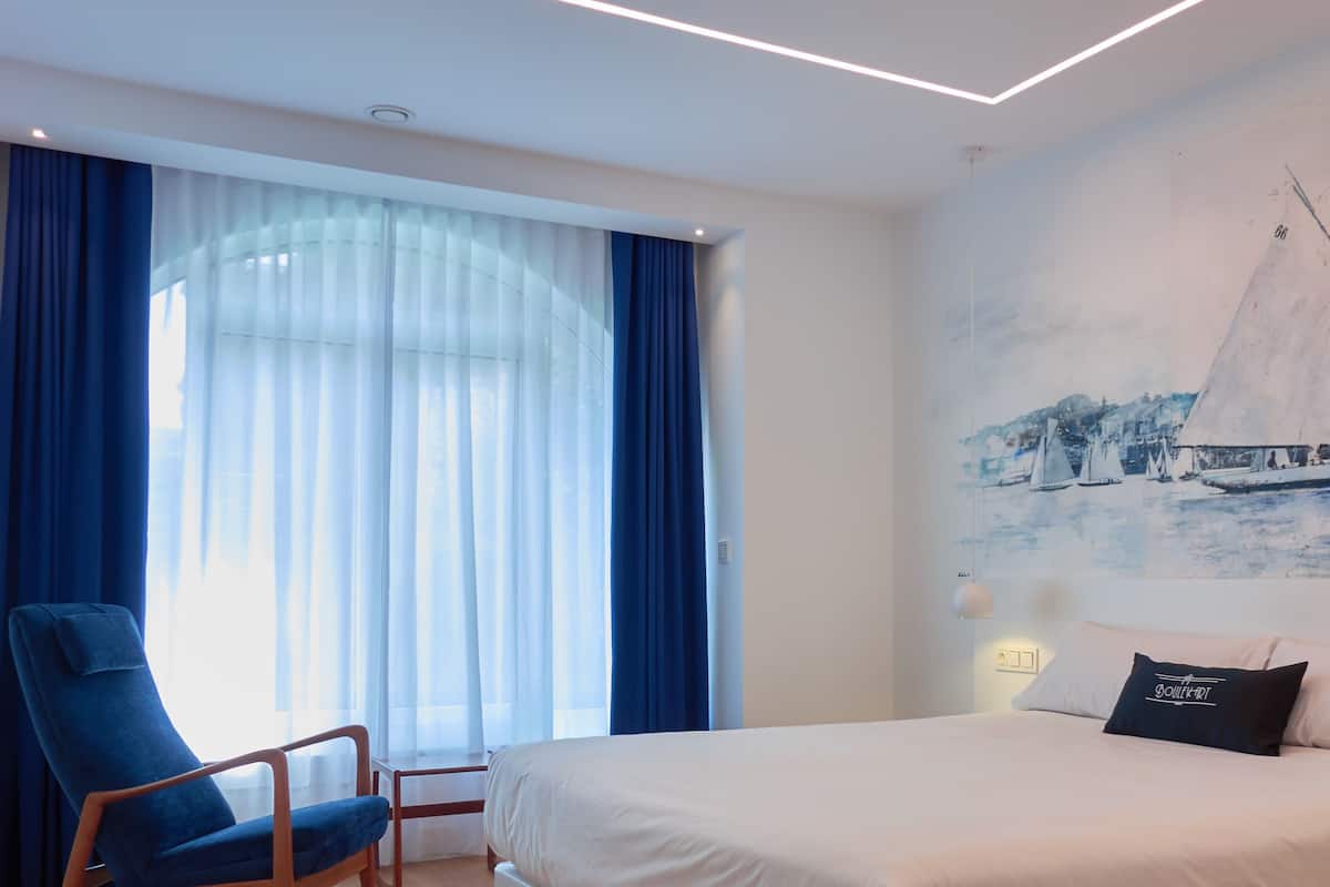 A hotel room with a bed, a chair, a painting of sailboats, and a window with curtains.