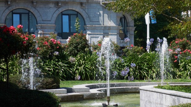 A fountain surrounded by lush greenery and vibrant flowers in front of a classical building with arched windows. - BoulevArt Donostia (San Sebastián)