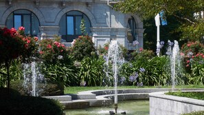 Une fontaine entourée d’une végétation luxuriante et de fleurs aux couleurs vives, située devant un bâtiment classique doté d’arcatures.