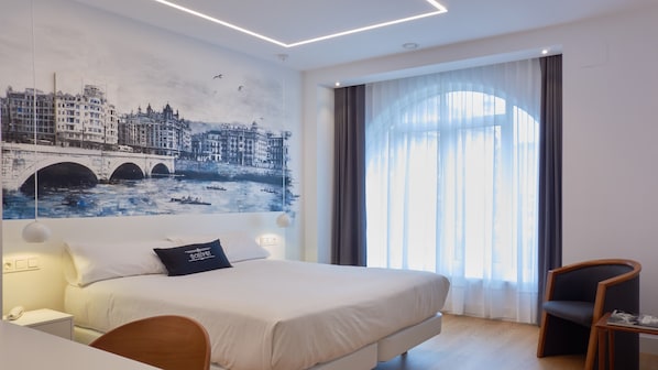 Una habitación de hotel moderna con una cama grande, un mural de paisaje urbano en la pared, un ventanal con cortinas y una silla de madera.