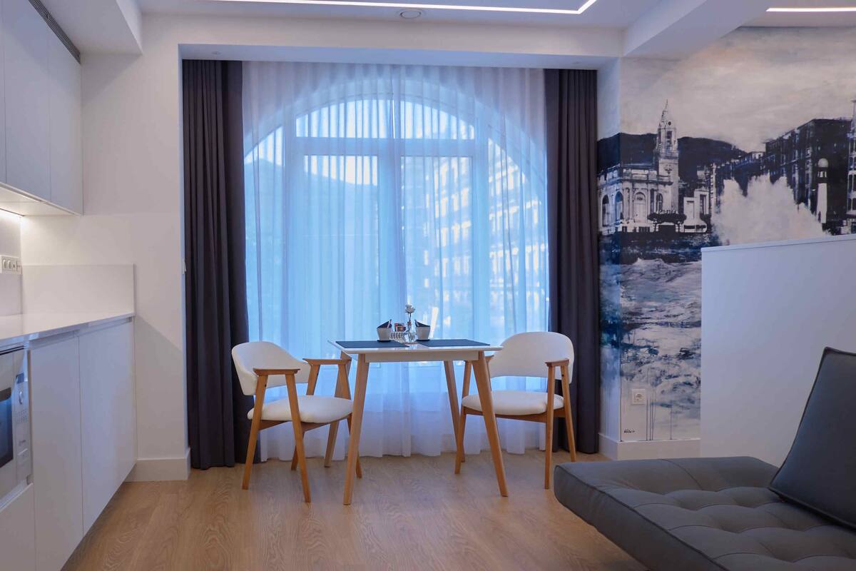A modern kitchen with white cabinets, a dining area with a table and chairs, and a large window with sheer curtains.