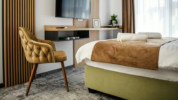A hotel room with a bed, a desk with a TV, a chair, and a window with curtains. - GBU Home - Olive Room (Timișoara)