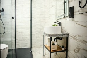 Uma casa de banho moderna com cabine de duche em vidro, lavatório branco sobre bancada de granito e um pequeno prateleira com uma planta.
