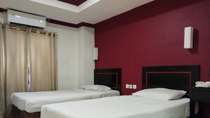 A hotel room with two single beds, a red accent wall, a ceiling fan, and an air conditioning unit.