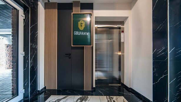 Um corredor moderno com uma placa na parede que diz “GBUHome”, um elevador de vidro e piso de mármore.