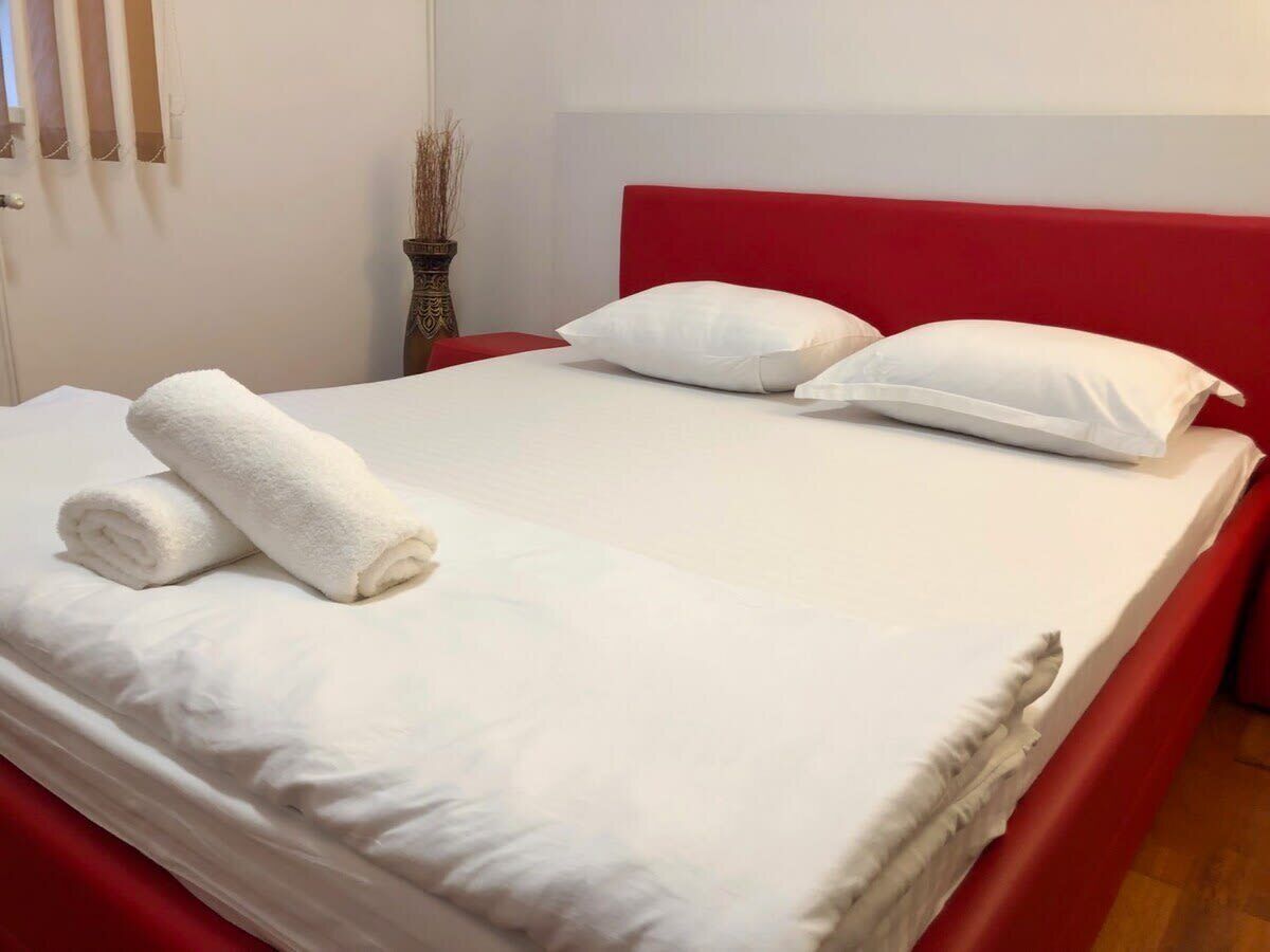 Ein ordentlich gemachtes Bett mit weißer Bettwäsche und einem zusammengerollten Handtuch, ein roter Bettrahmen und eine Vase mit getrockneten Zweigen auf dem Nachttisch.