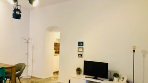Una sala moderna con suelo de madera, un televisor de pantalla plana sobre un soporte blanco, una mesa de comedor con sillas y una lámpara de pie.