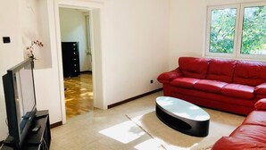 Un salon avec un canapé rouge, une table basse, un téléviseur posé sur un meuble et une vue sur la verdure par la fenêtre.