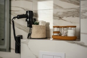 Casa de banho com parede de mármore, secador de cabelo preto, cesto de fibra natural e bandeja com frascos e uma planta.