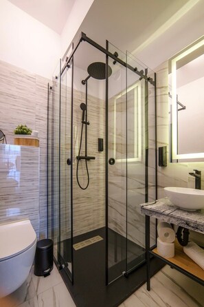 A modern bathroom with a glass-enclosed shower, a marble-tiled wall, a white sink, and a large mirror.