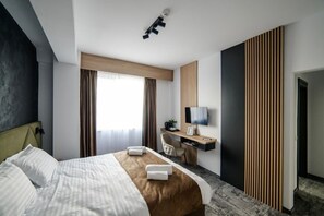 A modern hotel room with a large bed, a desk with a chair, a flat-screen TV, and a large window with curtains.