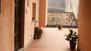 A corridor with a tiled floor, a black door, a wall-mounted light fixture, and a view of a terracotta-tiled balcony with potted plants. - I Borghi della Maddalena (Borgo a Mozzano)