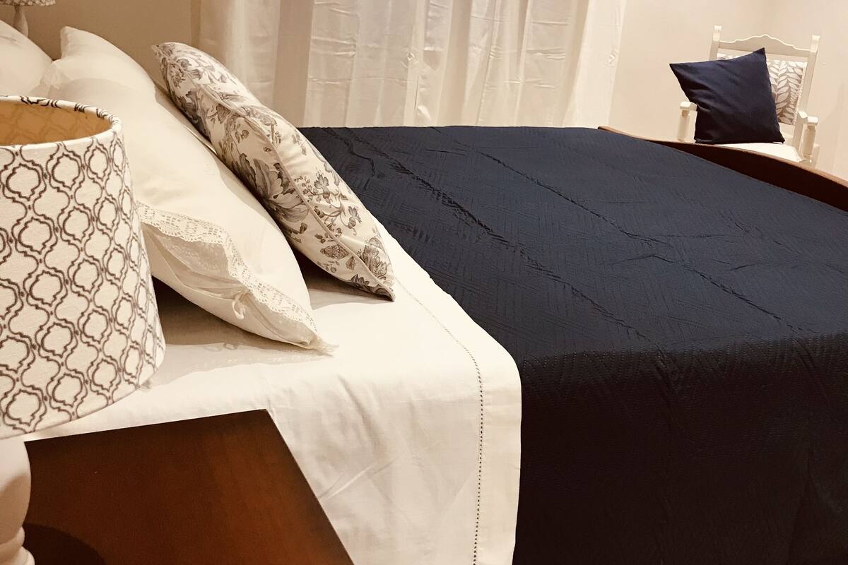 A neatly made bed with a dark blue bedspread and white linens, a wooden nightstand with a lamp, and a chair with a blue cushion.