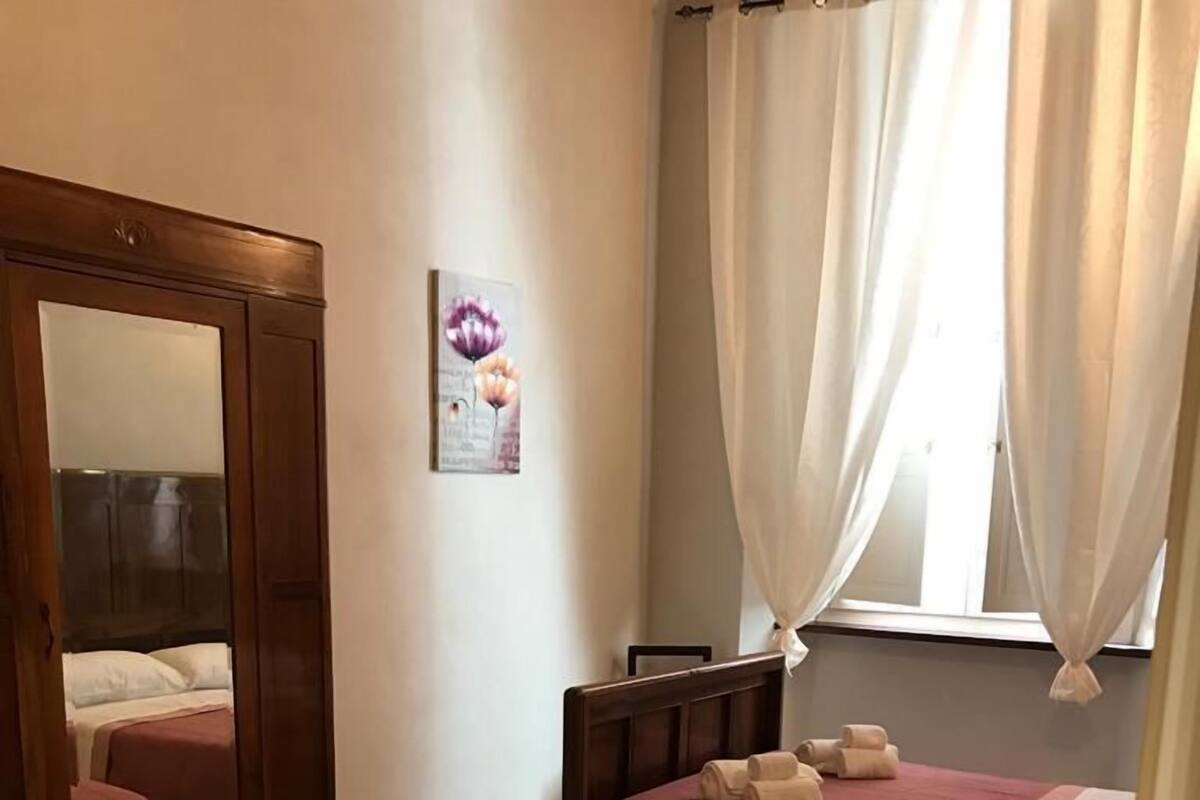A hotel room with two beds, each with a wooden headboard and a red bedspread. There is a large mirror on the left wall and a window with white curtains.