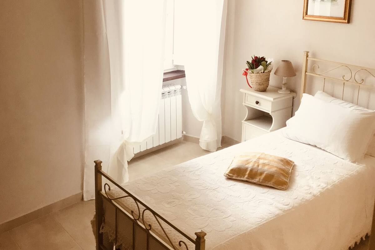 A bedroom with a bed, a nightstand, a lamp, a framed picture, and a window with curtains.