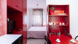 A compact room with a bed, a television, a desk, and a dining area with a red cabinet and a potted plant.