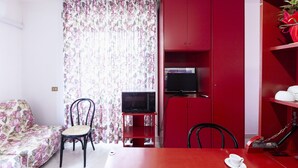 A compact living space with a floral-patterned sofa, a dining table with a cup, a red cabinet with a television, and a chair.