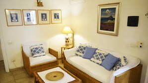 A cozy room with a sofa, two armchairs, a small table with a mat, a lamp, and framed pictures on the wall. - Beachfront vacation home in elegant residential complex near Chia (Pula)
