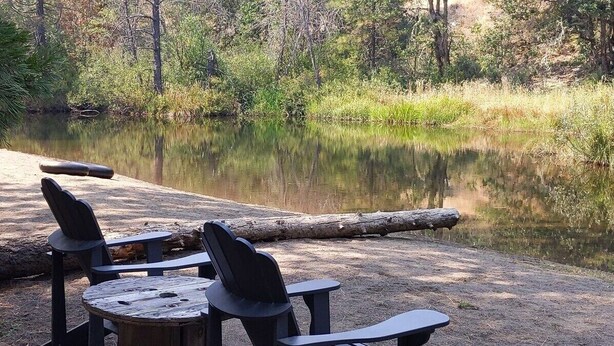 Ein Campingplatz mit Picknicktischen und -stühlen an einem ruhigen See, umgeben von Bäumen.