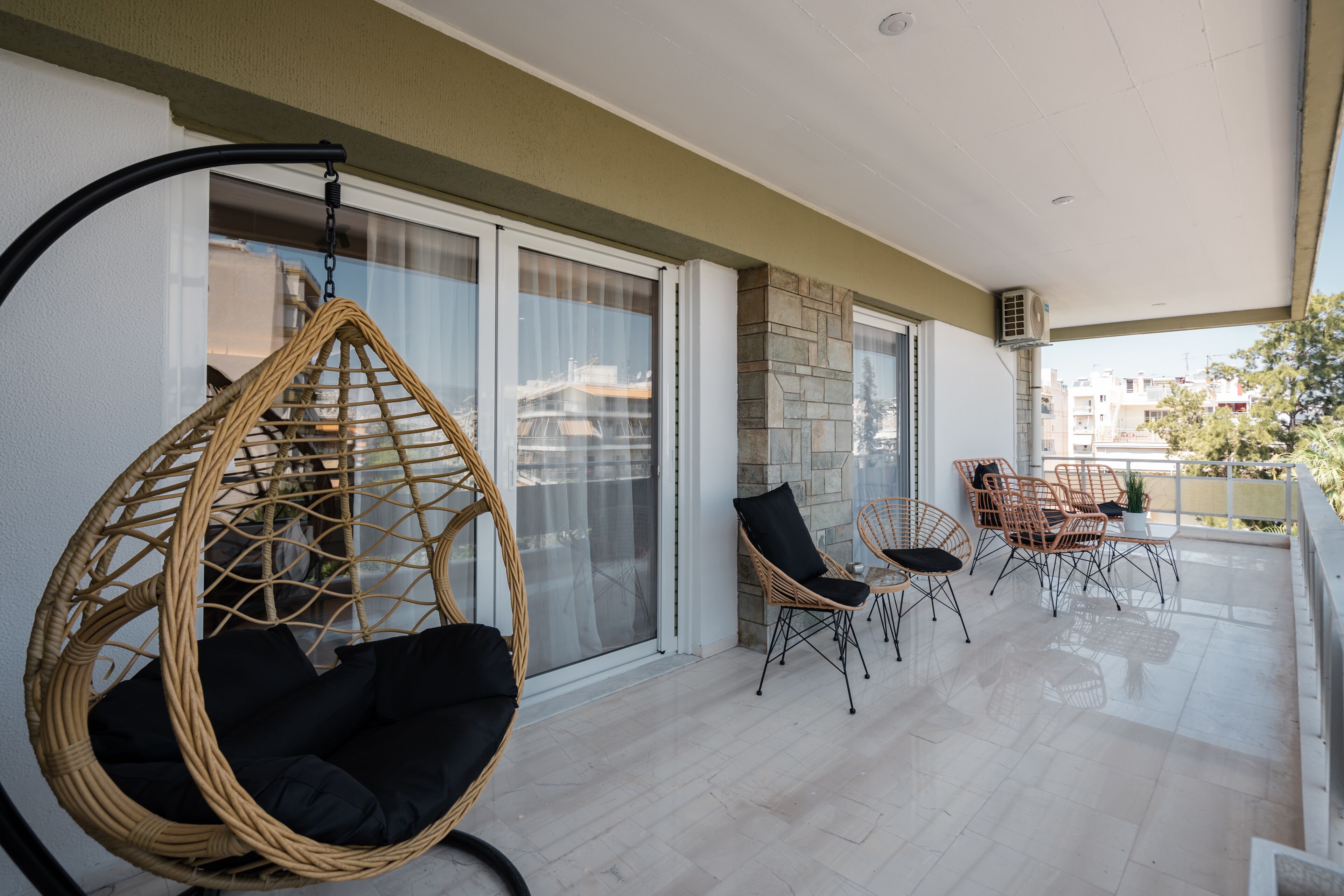 A balcony with wicker chairs and a hanging chair, large windows, and a tiled floor.