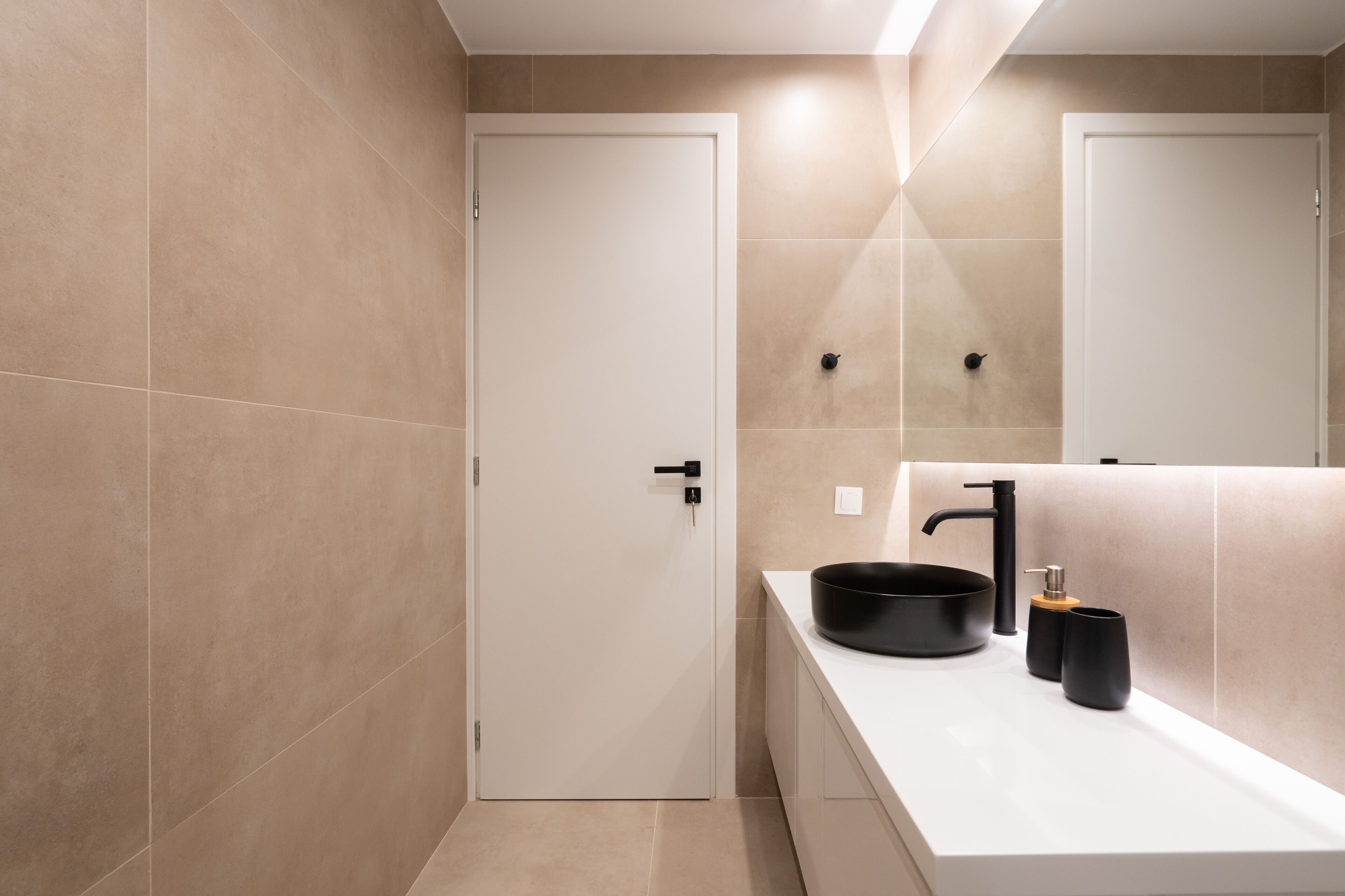 Une salle de bain moderne avec des carreaux de couleur beige, un meuble-lavabo blanc, un évier noir et un miroir.