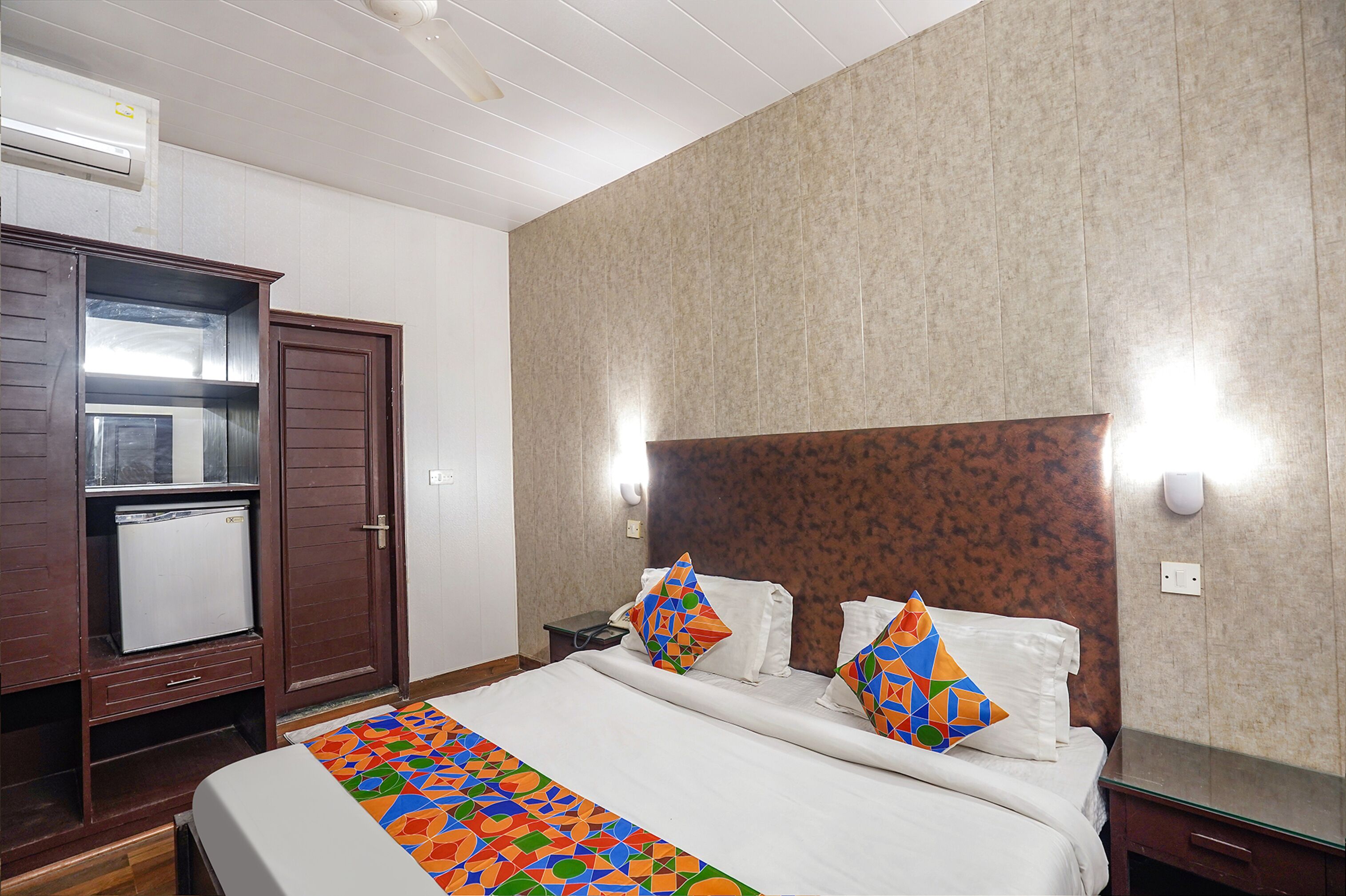 A hotel room with a bed, a small refrigerator, a wooden wardrobe, and a tiled wall.