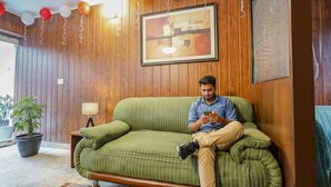 Ein Mann sitzt auf einem grünen Sofa in einem Raum mit Holzvertäfelung, einem gerahmten Bild an der Wand und Ballons.