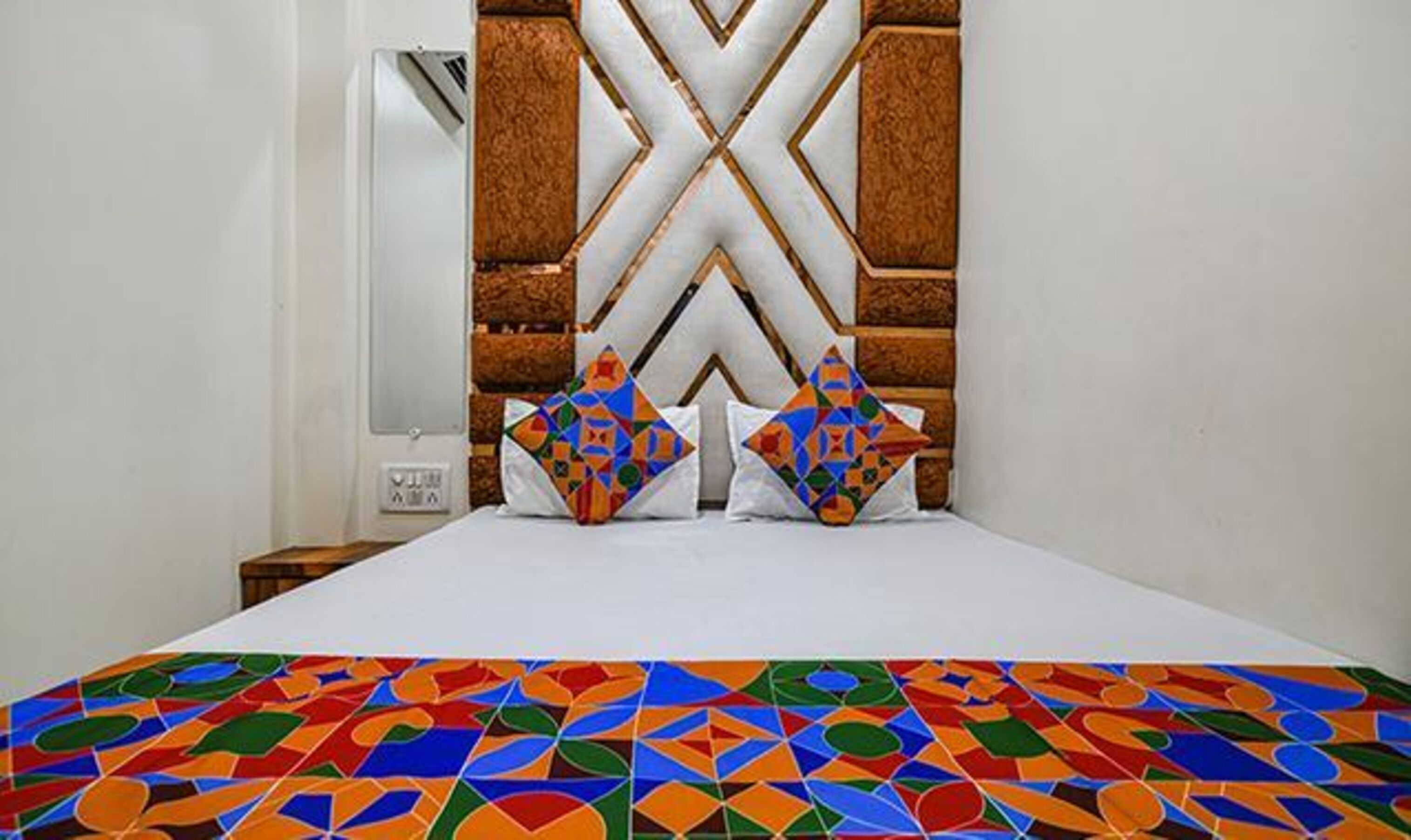 A hotel room with a bed, a headboard with a geometric pattern, and a colorful geometric-patterned bedspread.