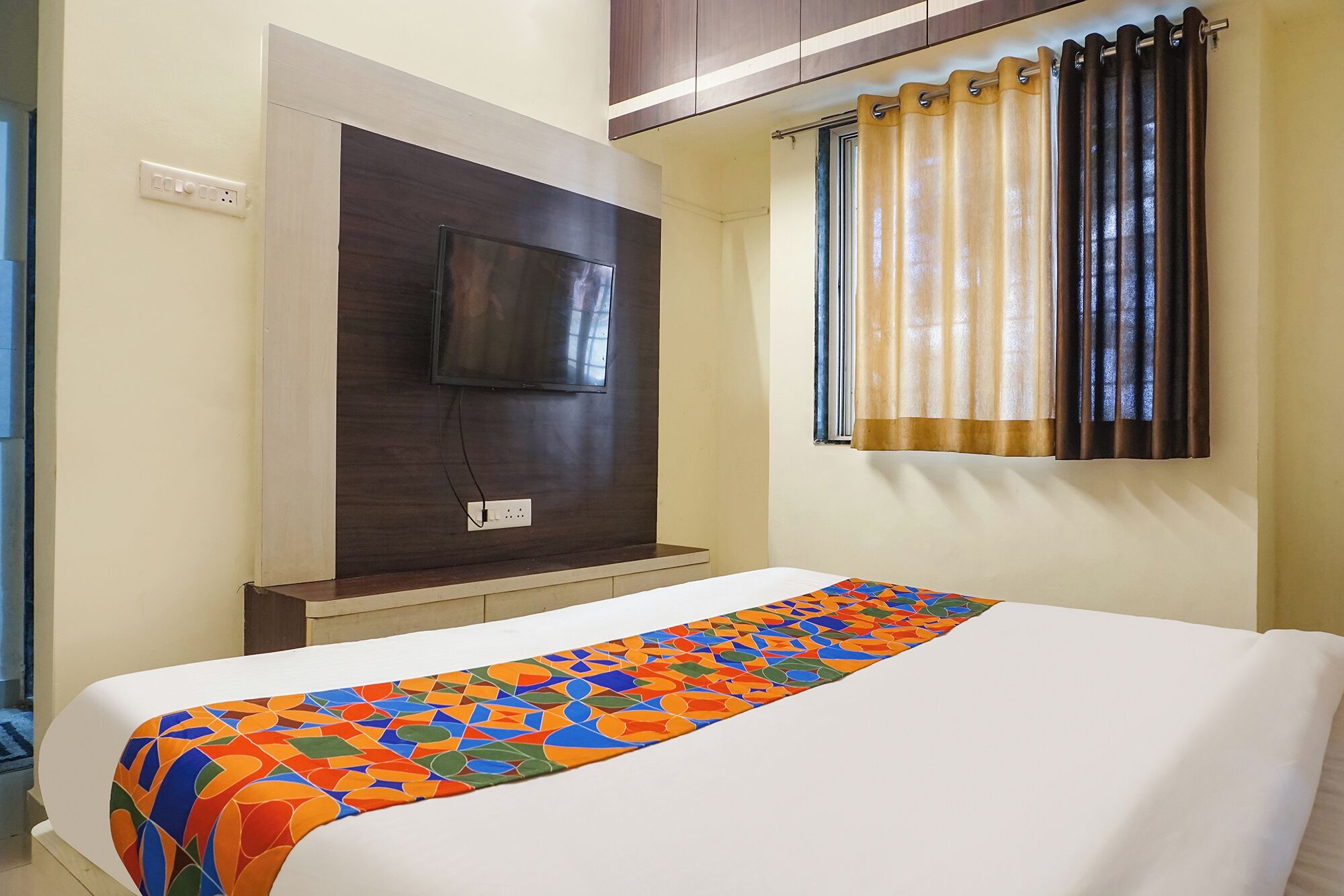 A hotel room with a bed, a TV mounted on a dark wooden panel, and a window with beige curtains.