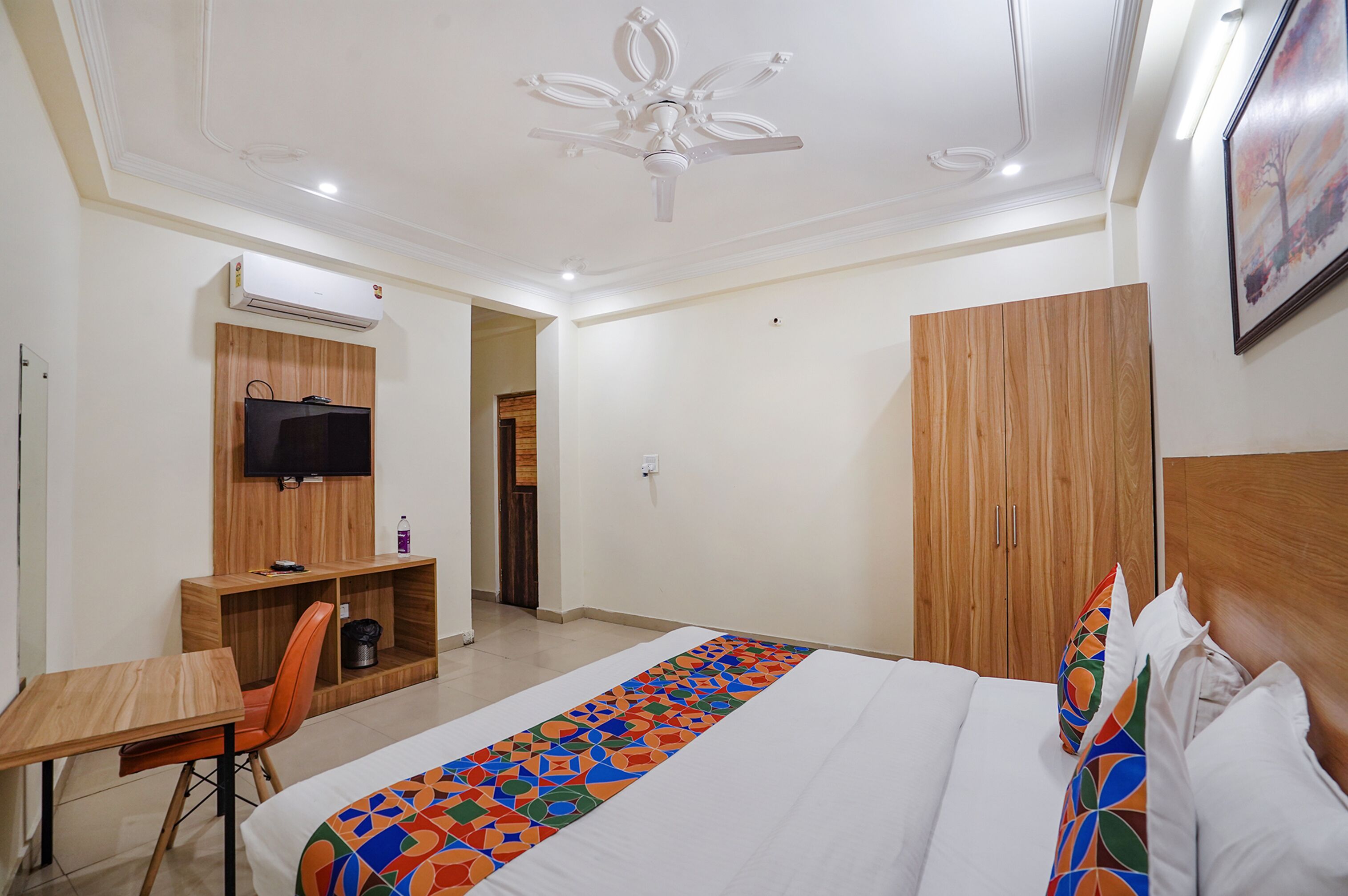 A hotel room with a bed, a desk with a chair, a television, a wardrobe, and a ceiling fan.