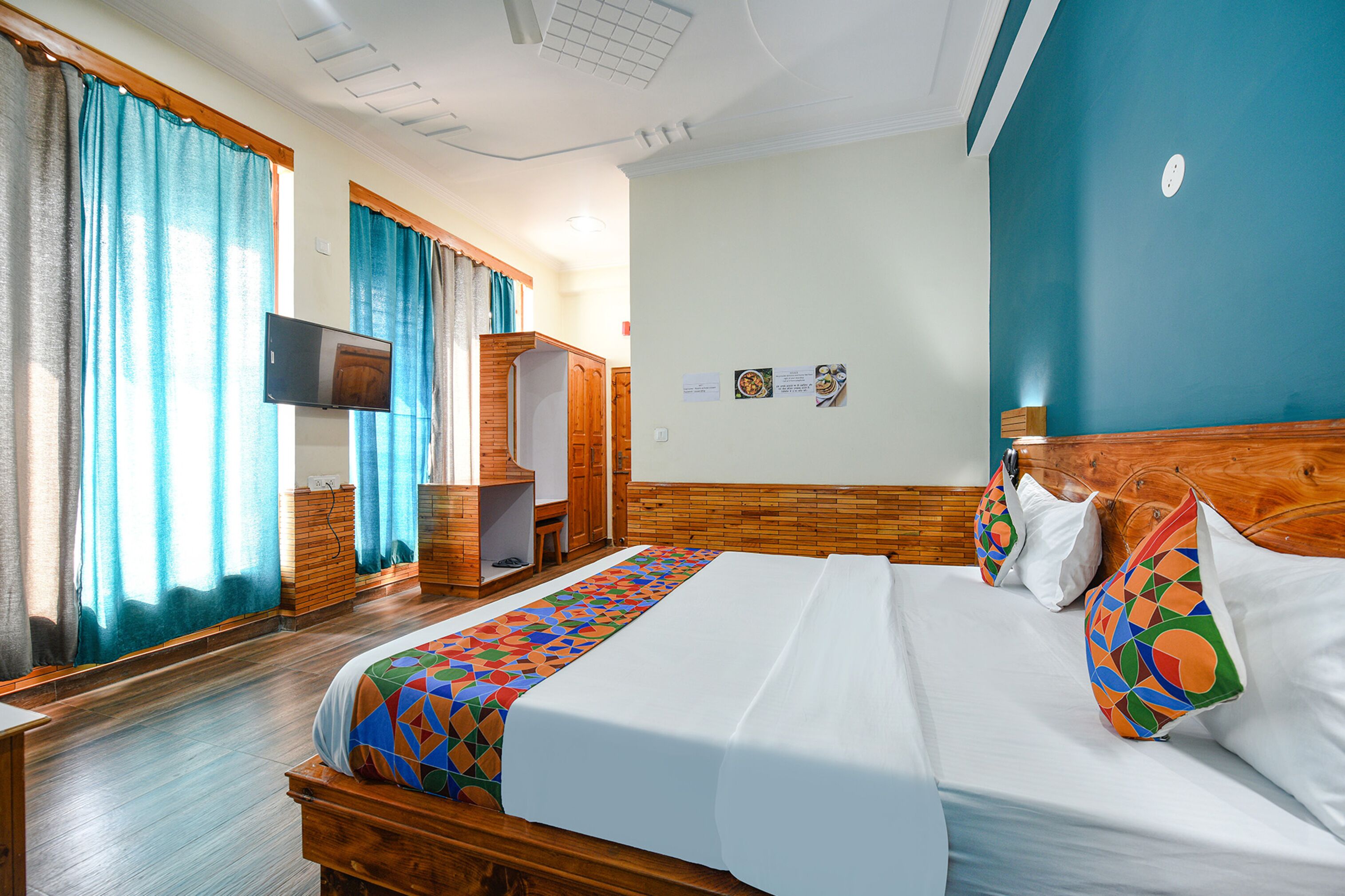 A hotel room with a large bed, wooden headboard, a television on the wall, and a window with curtains.