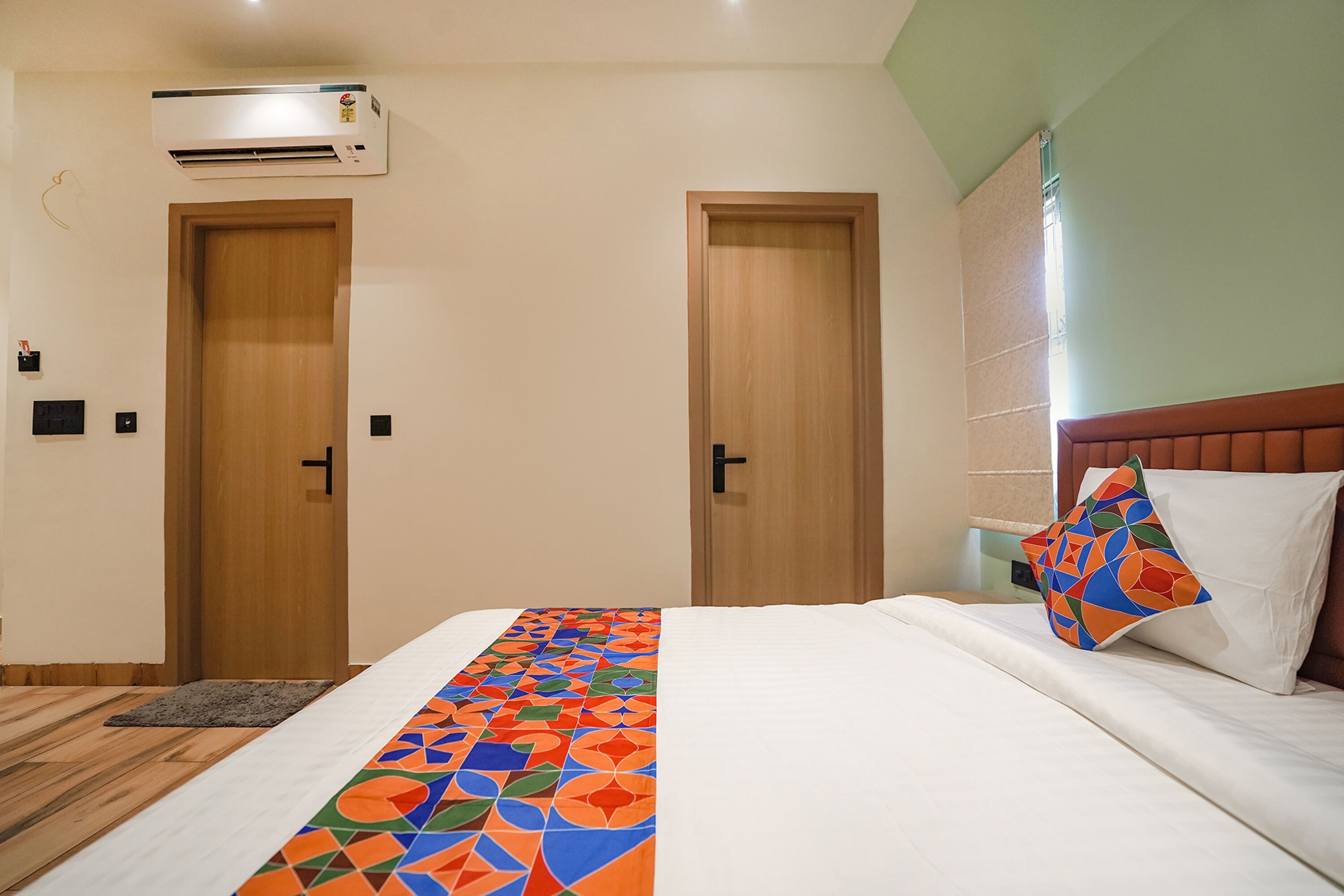 Habitación de hotel con cama, un corredor de cama de estampado geométrico colorido, dos puertas de madera, un almohadón blanco y un aire acondicionado instalado en la pared.