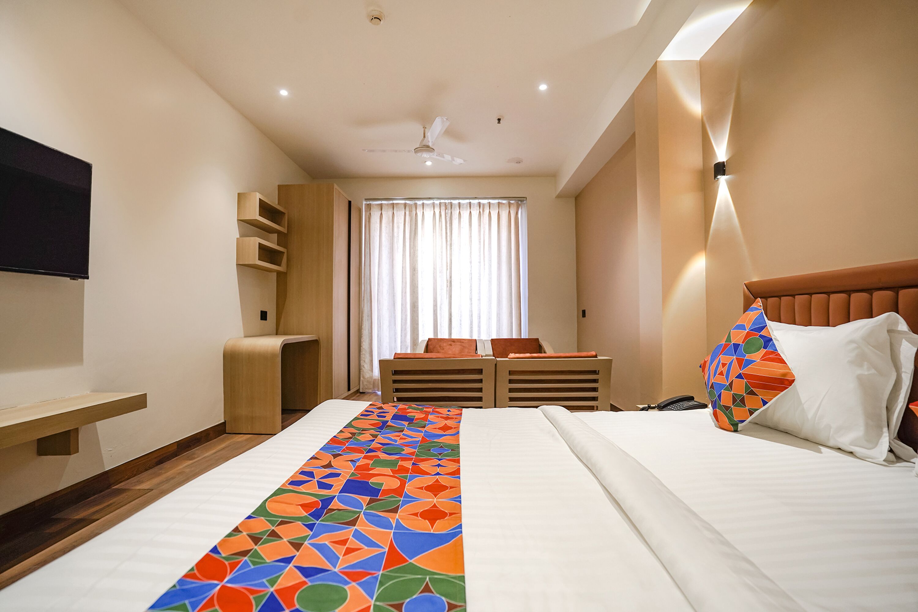 A hotel room with a bed, a TV mounted on the wall, a desk with a chair, and a window with curtains.