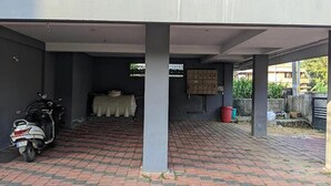 A covered parking area with a scooter parked on the left, a brick floor, and a covered seating area with a table and chairs in the center. - Fabexpress Aabriz Residency (Kanayannur)