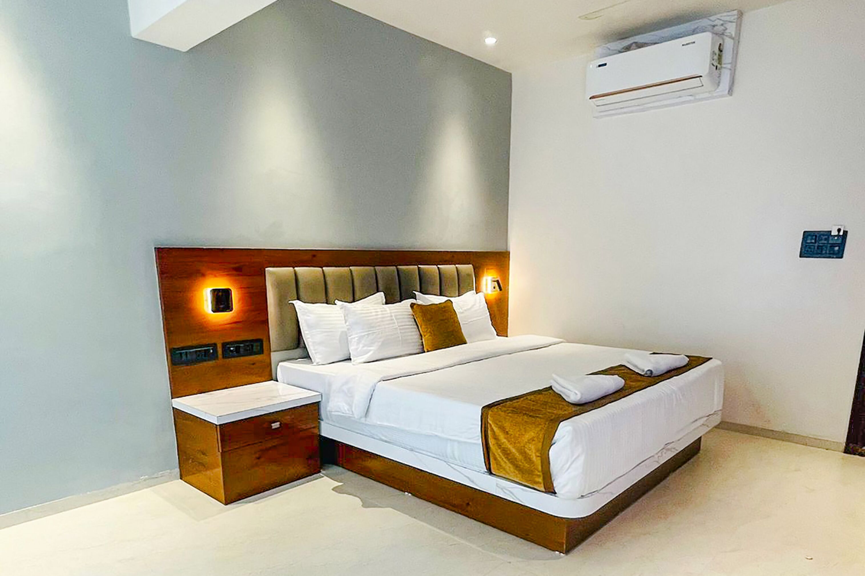 A modern hotel room with a large bed, bedside tables, a built-in nightstand, and a wall-mounted air conditioning unit.
