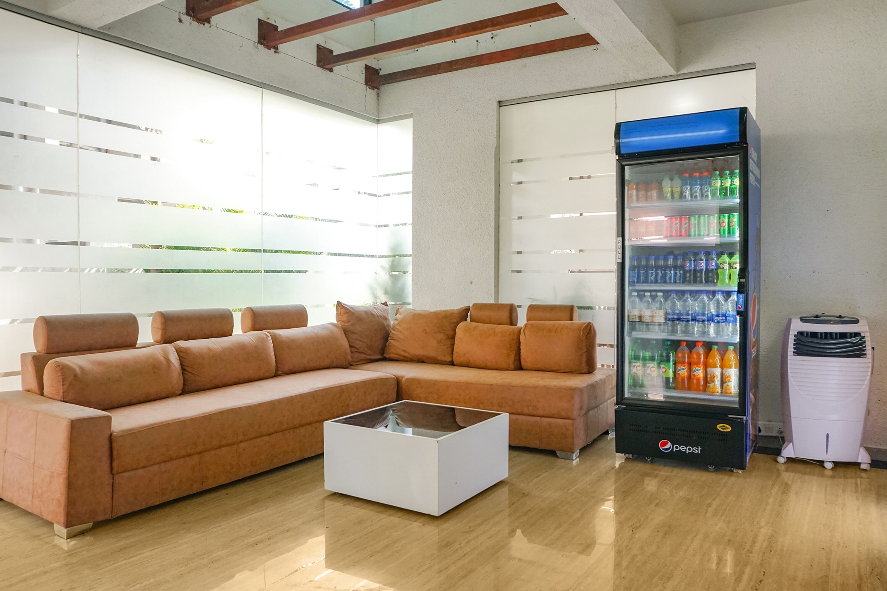 Un espace de détente moderne comprenant un canapé d’angle, une table basse en verre, un réfrigérateur Pepsi et un climatiseur portable.