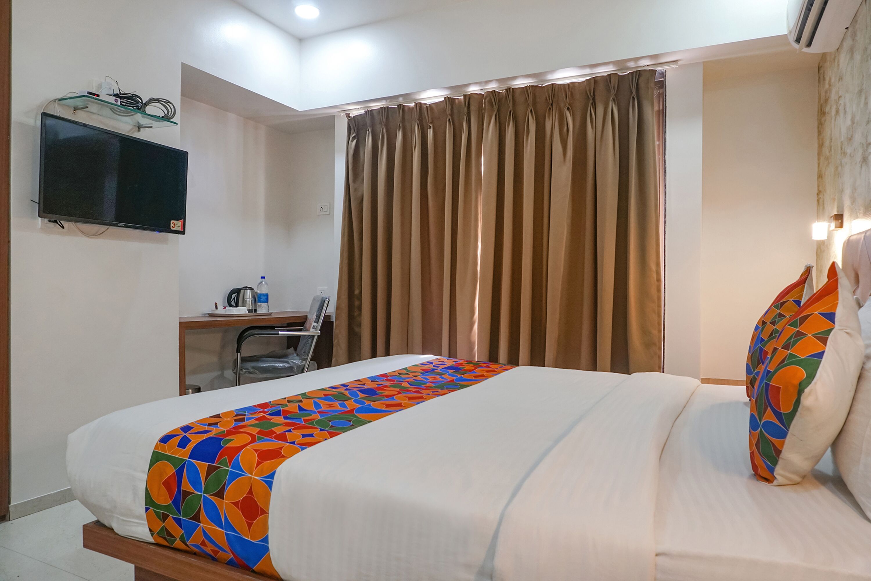 Une chambre d’hôtel comprenant un lit, un bureau avec une chaise, un téléviseur fixé au mur et une fenêtre avec des rideaux.