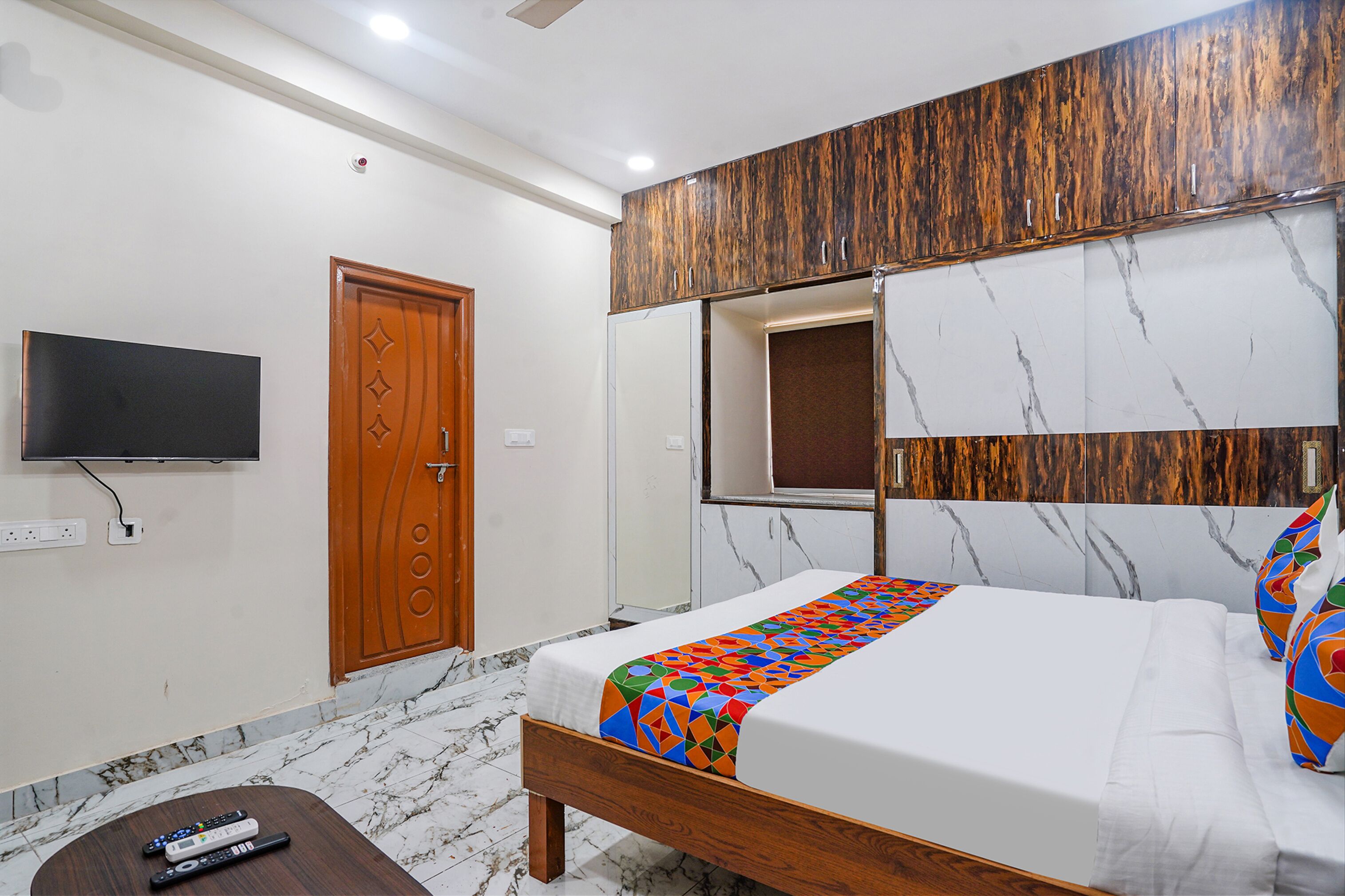 Un dormitorio con cama, televisor montado en la pared, puerta de madera y suelo de mármol.