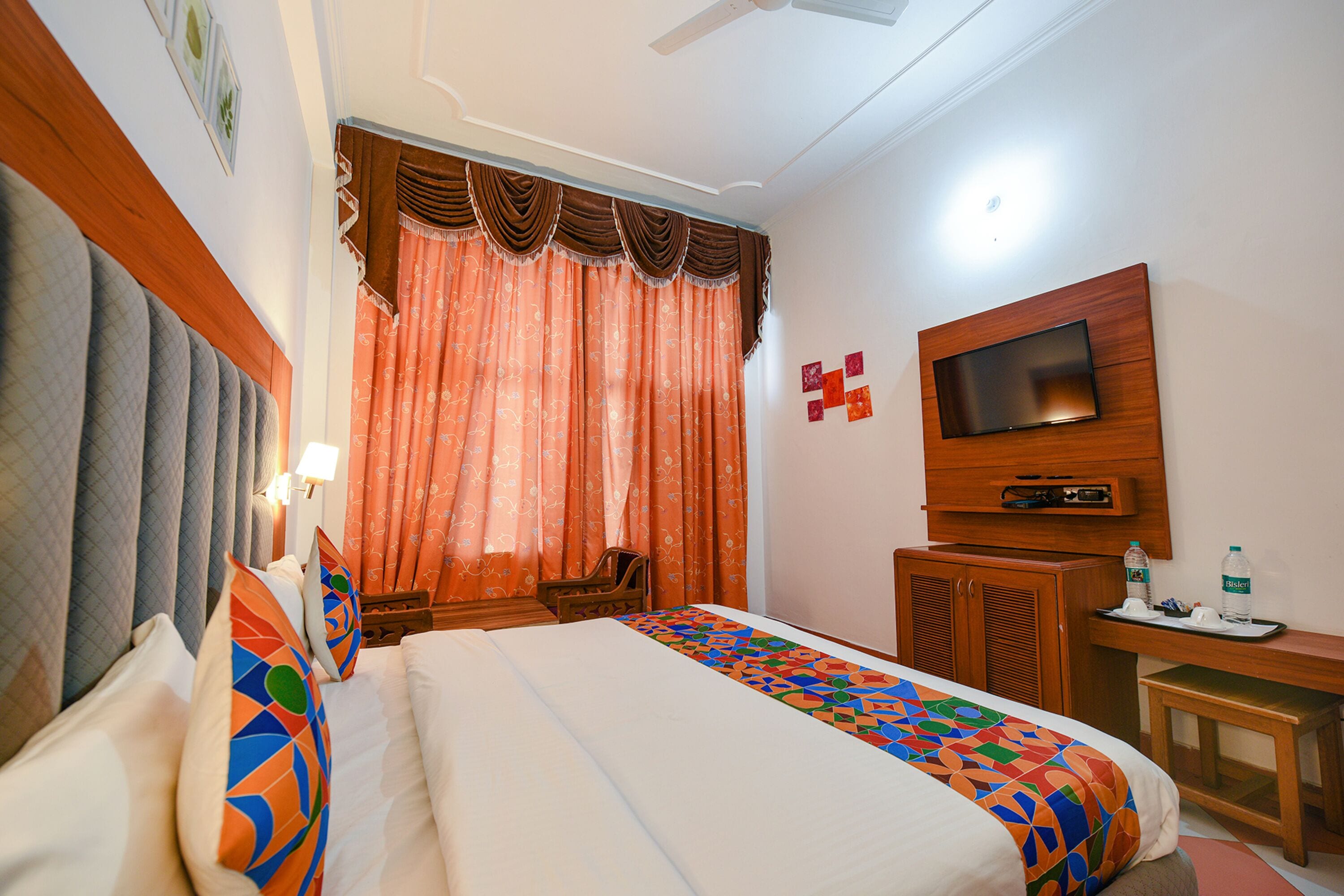 A hotel room with a bed, a TV mounted on the wall, a desk with a chair, and a window with curtains.