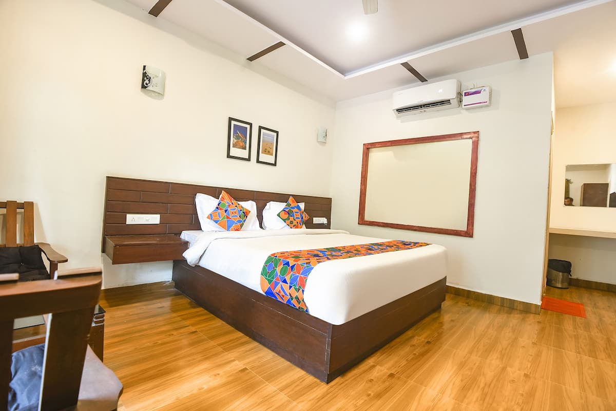 A hotel room with a wooden floor, a bed with a colorful geometric bedspread, a wooden headboard, a large mirror, and an air conditioning unit.