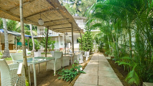 An outdoor seating area with wicker furniture under a shaded structure, surrounded by lush greenery and a paved walkway. - Fabhotel La Volga (Morjim)