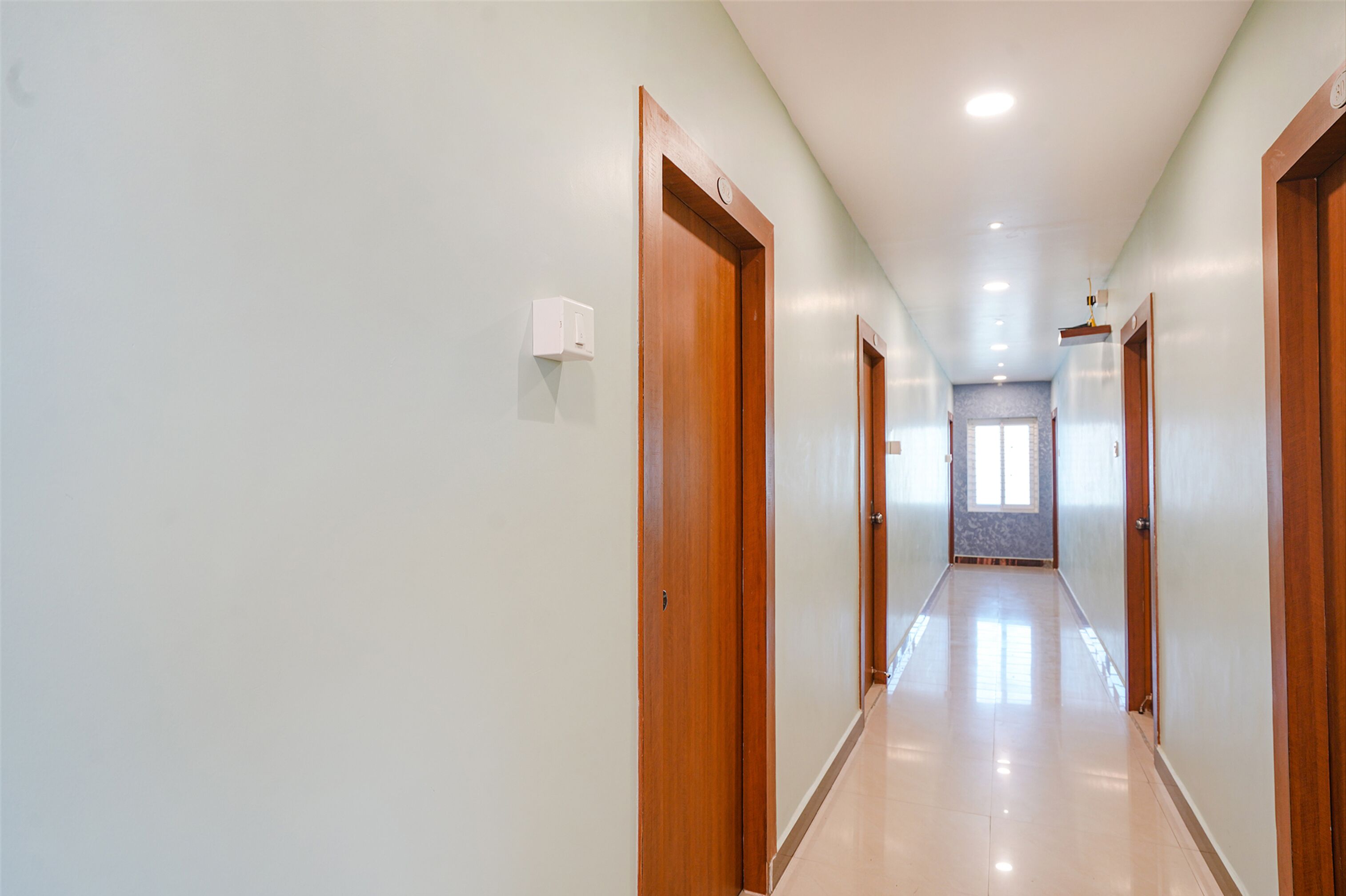 A long, narrow hallway with wooden doors on both sides, leading to a room with a large window at the end.